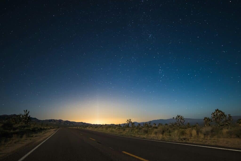 Desert highway stretching into the distance under a star-filled night sky, with a faint glow on the horizon.