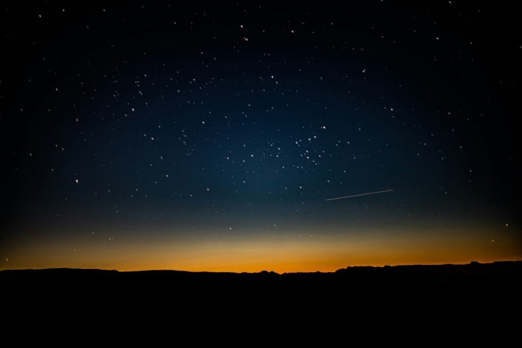 Starry night sky over a silhouetted desert landscape, with a faint meteor streaking across the horizon.