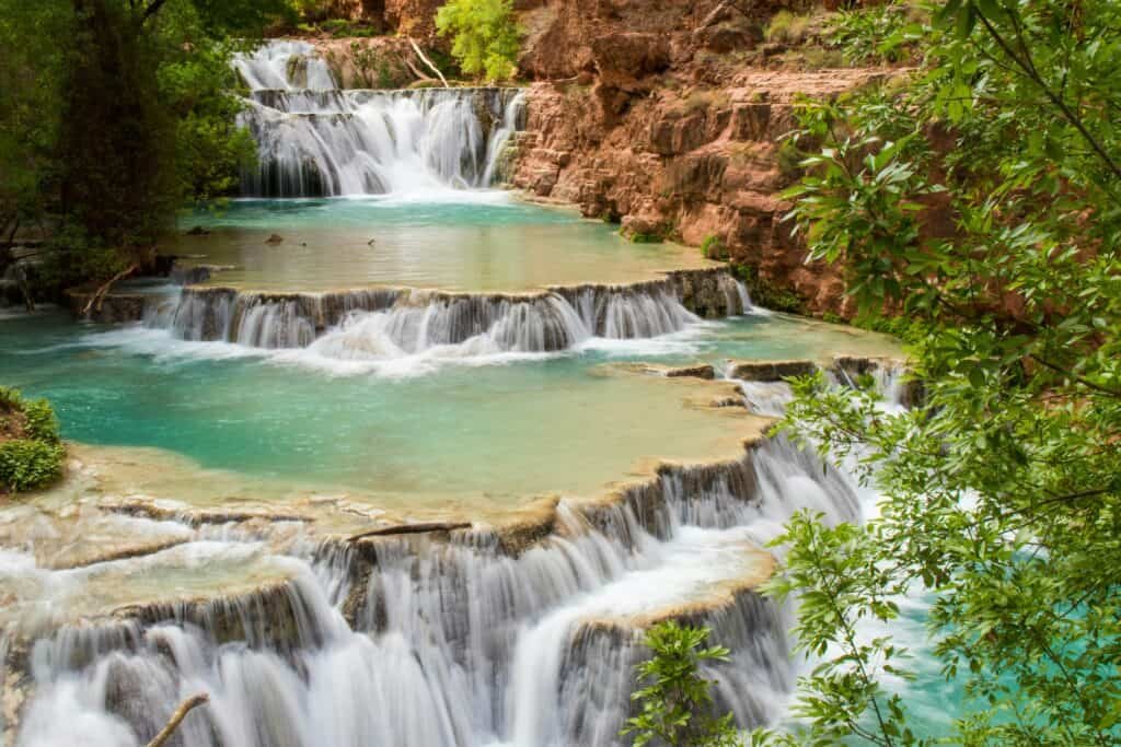 Cascading turquoise waterfalls flowing over tiered rock formations, surrounded by lush green foliage.