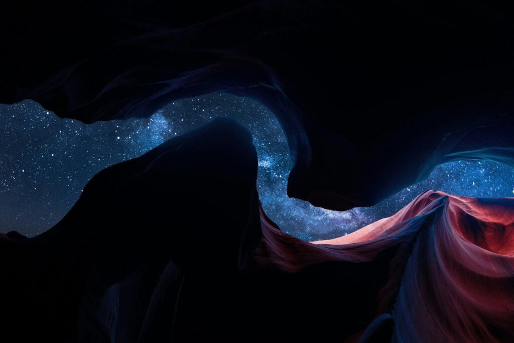 View of the Milky Way through the curves of a slot canyon, with the sandstone walls illuminated by a soft glow.