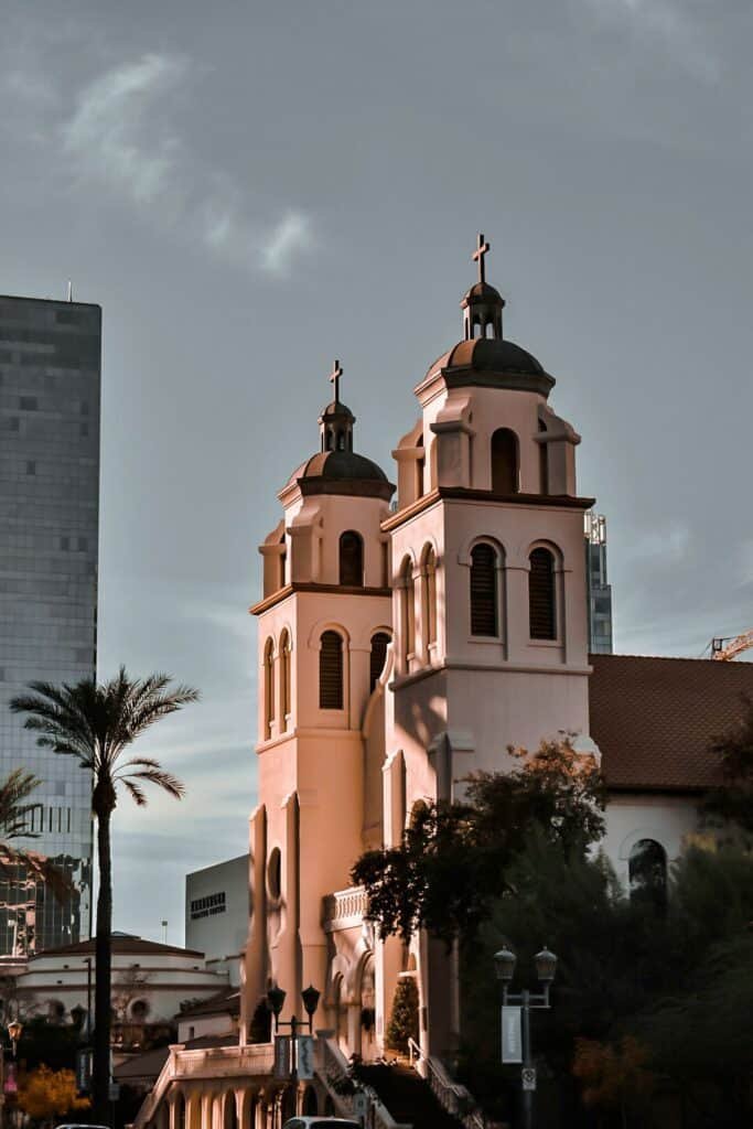 Historic church with twin bell towers illuminated by warm sunlight, set against the Tucson cityscape with palm trees and high-rise buildings.