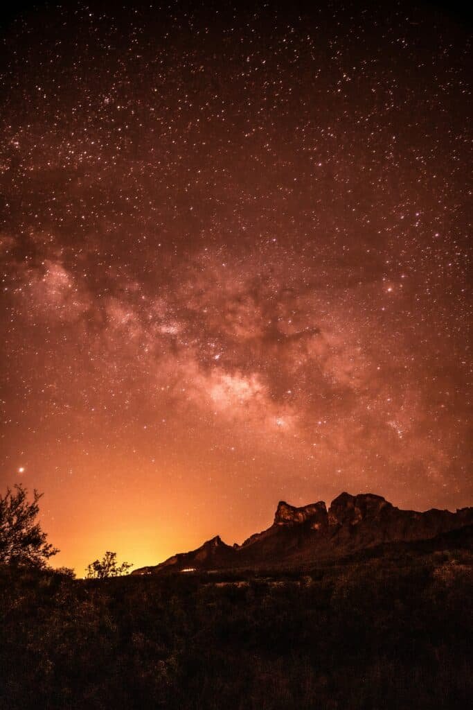 Night sky over a rugged mountain range with the Milky Way galaxy visible, glowing against a warm, orange-hued horizon.